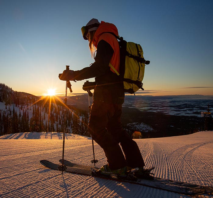 A skiier stands at a hill, with the sun setting in the horizon.