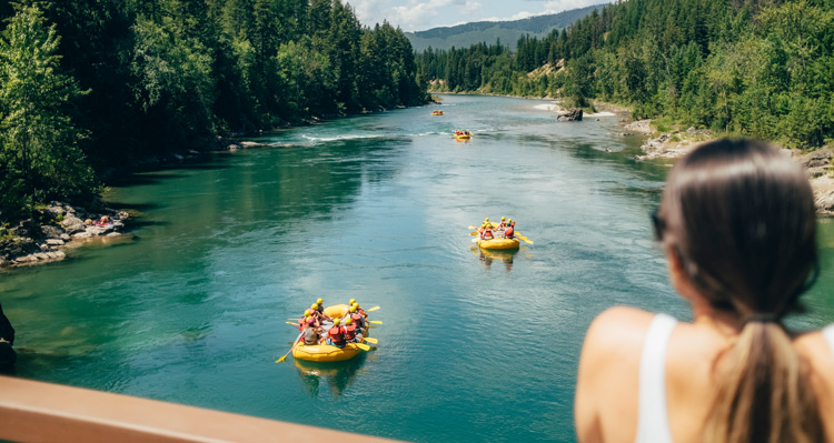 A person standing on a bridge watching rafting groups float down the Flathead River towards the mountains.
