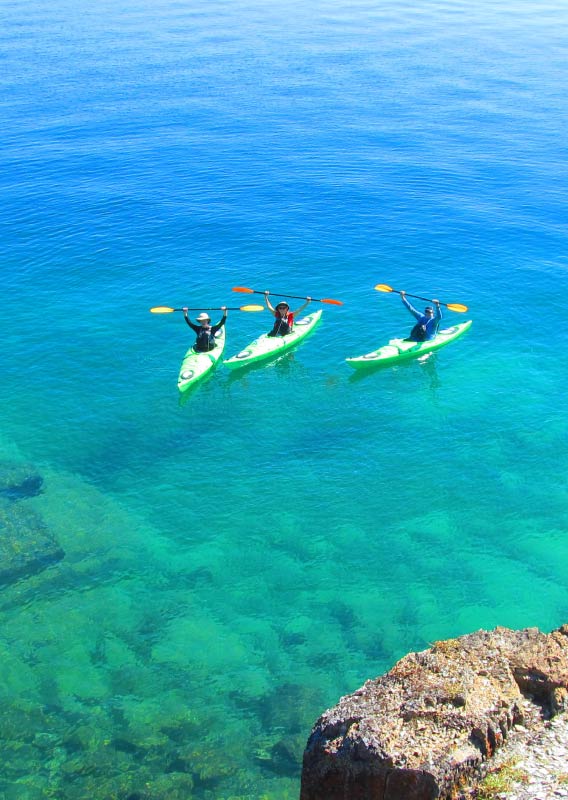 Three kayakers raise their paddles over their heads as they float on a clear blue lake.