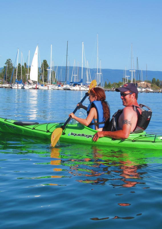 A man and a child sit in a kayak, paddling on a lake near a marina.