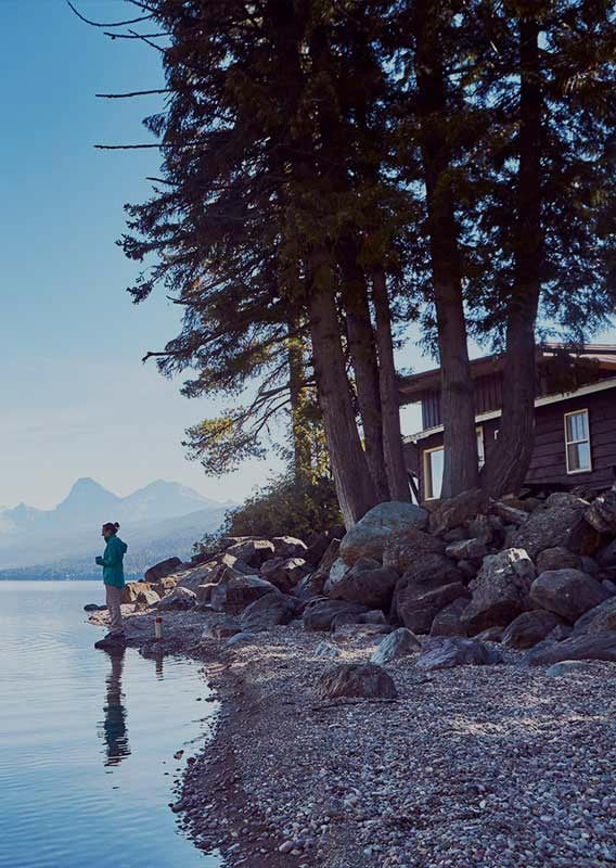 Woman stands on bank of calm Lake McDonald as a man in a canoe approaches