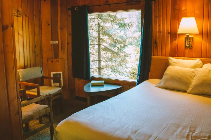 A bed sits next to a large window in a wooden cabin.