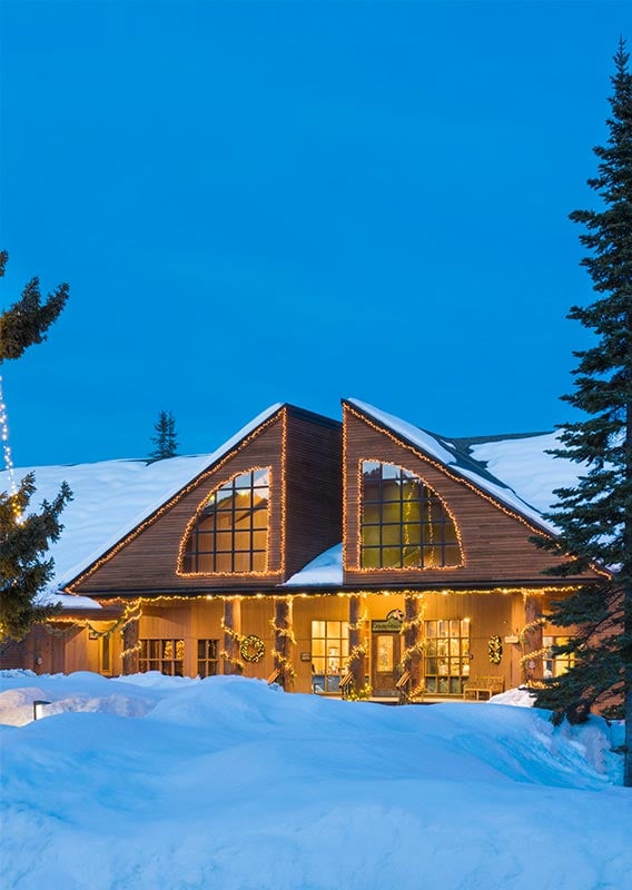 A lodge with large windows covered with snow and Christmas lights.