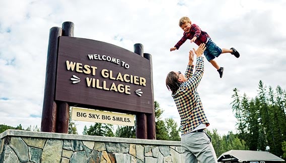 A dad raises a child above in front of the entry sign to West Glacier Village