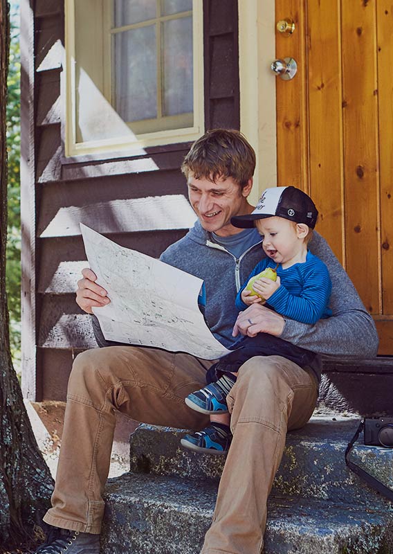 A dad sits with a child on his lap on the steps of a wooden cabin