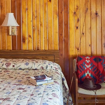 Interior of a wooden lodge room, with a bed, chair and fishing pole against the wall.