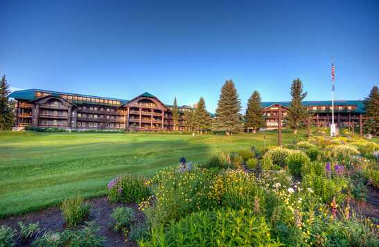 A large wooden lodge behind a wide green lawn and gardens.