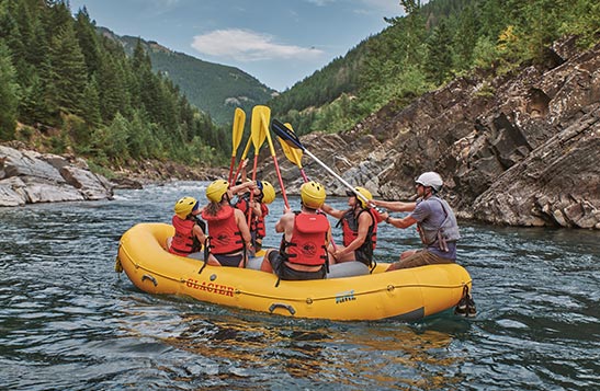 A group of rafters on a boat with kids lift their paddles to touch the tips.
