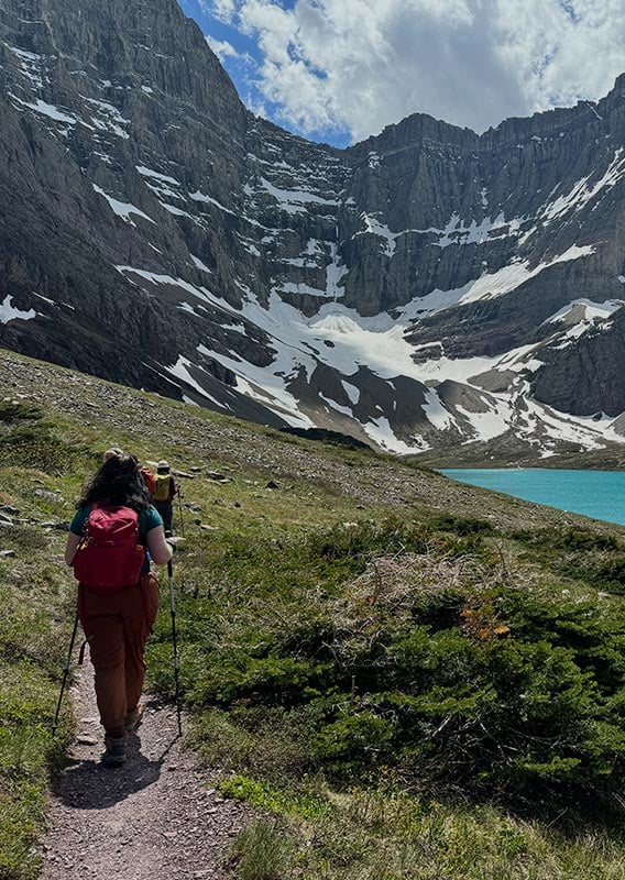 A hiker walks on a trail above a blue lake towards tall mountains.